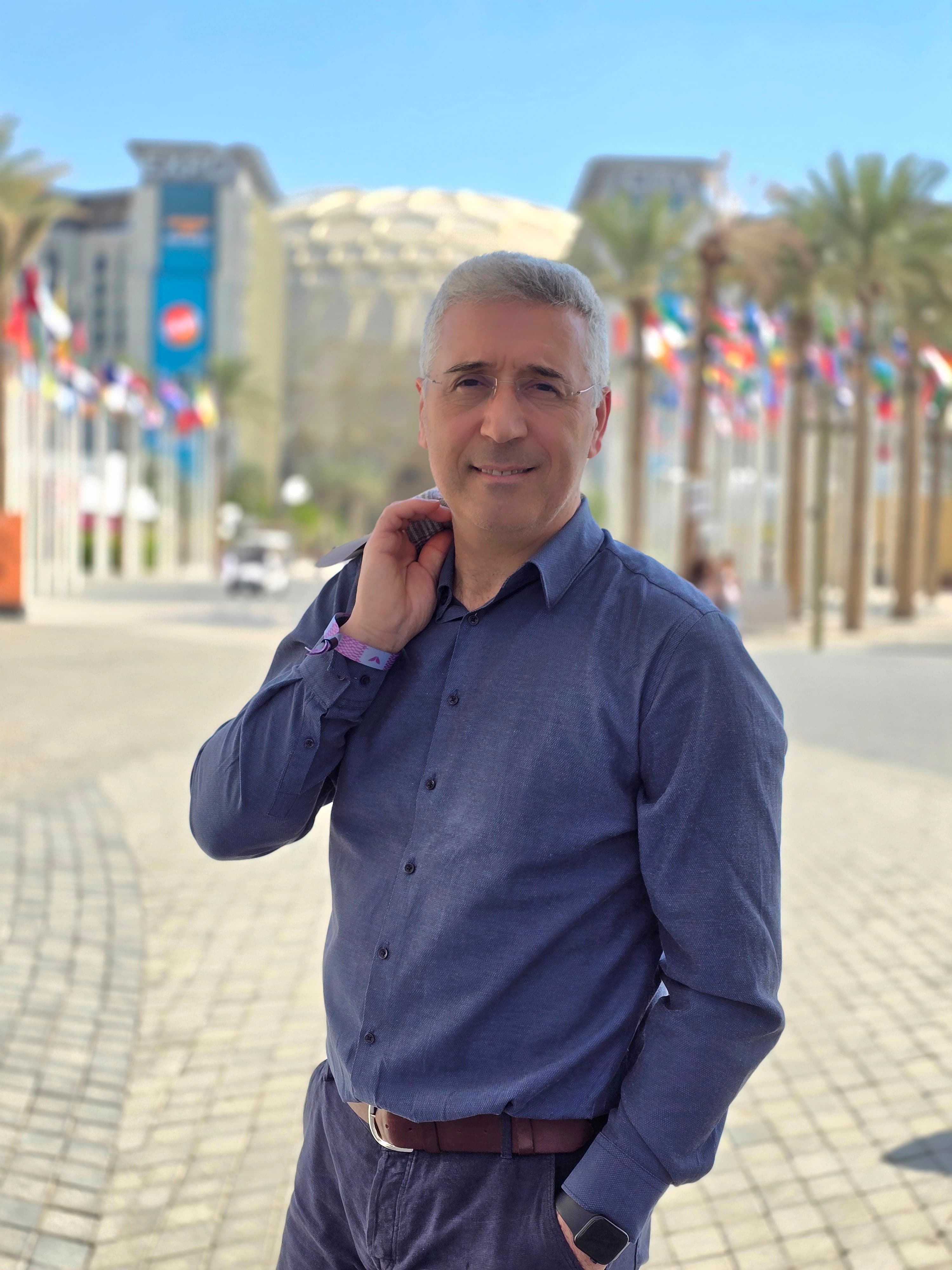 Smiling man with glasses in a blue shirt holding a jacket outdoors with international flags.